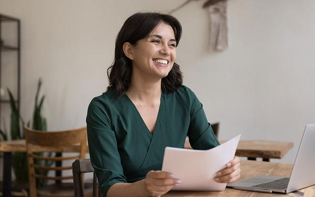 Junge Frau mit strahlendem Lächeln sitzt am Schreibtisch und hält optimierte Bewerbungsunterlagen in der Hand, neben ihr ein Laptop für die Bewerbungsvorbereitung.