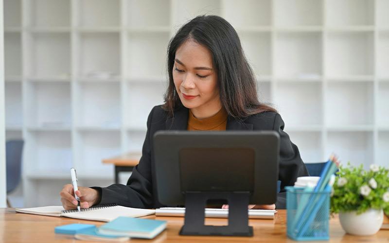 Frau sitzt im Büro vor einer riesigen Regalwand mit leeren Fächern und macht sich Notizen an ihrem Schreibtisch.