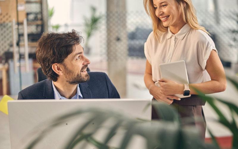 Mann im Büro am Schreibtisch und eine neben ihm stehende Frau unterhalten sich über das Thema Umschulung Büromanagement Teilzeit.