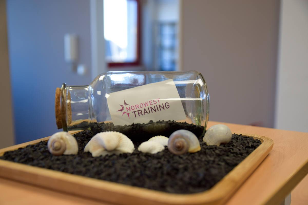 Kleine Flaschenpost mit einem beschrifteten Zettel von Nordwest Training darinnen auf dekorativer Unterlage mit Muscheln.